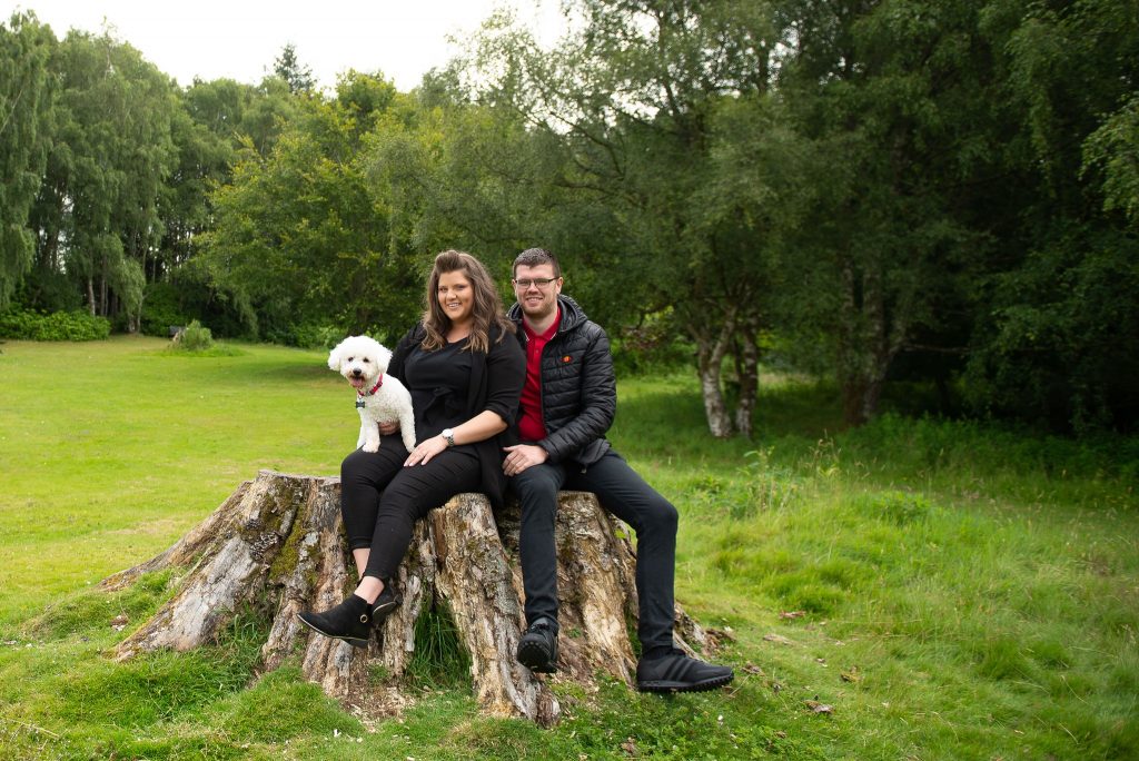 couple with dog sad on giant tree stump with large pine trees in the background.